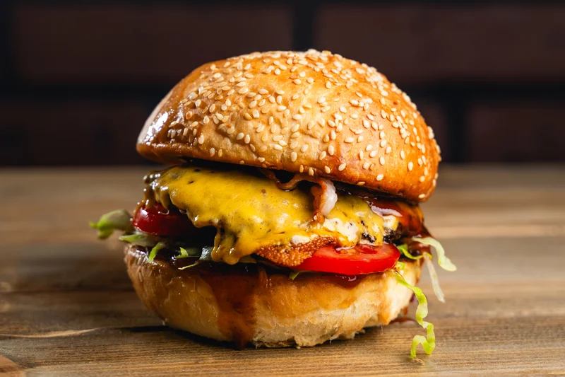 USA Cheese Burger 1 Szezámmagos kézműves hamburger, tökéletesre pirított baconnel, vörös cheddar sajttal, friss paradicsomkarikával és salátával, lilahagymával.